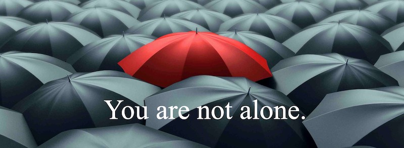 Opened red umbrella in the middle of a sea of opened grey umbrellas with texts saying “You are not alone.”